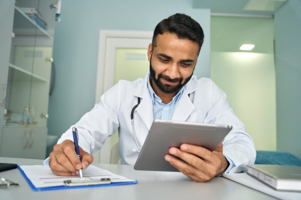 Indian happy male doctor in clinic using tablet device writing health data.